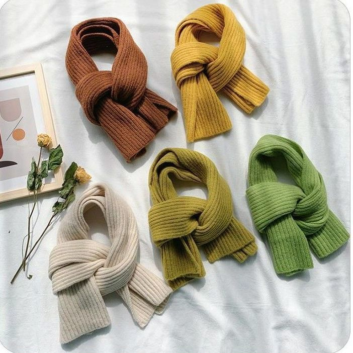 plusieurs écharpes du même modèle, présenté noué, à plat sur un tissu blanc , près de deux fleurs jaunes, il y a 5 couleurs différentes : marron, jaune, beige, kaki, et vert
