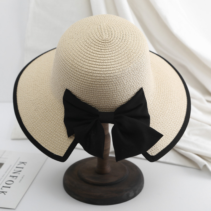 Chapeau de paille beige de dos sur une porte chapeau en bois avec gros nœud en tissu noir à l'arrière et un liseré de tissu sur le bord. Le chapeau à une ouverture sous le nœud. Le tout posé sur une table blanche avec un tissu blanc dans le fond.