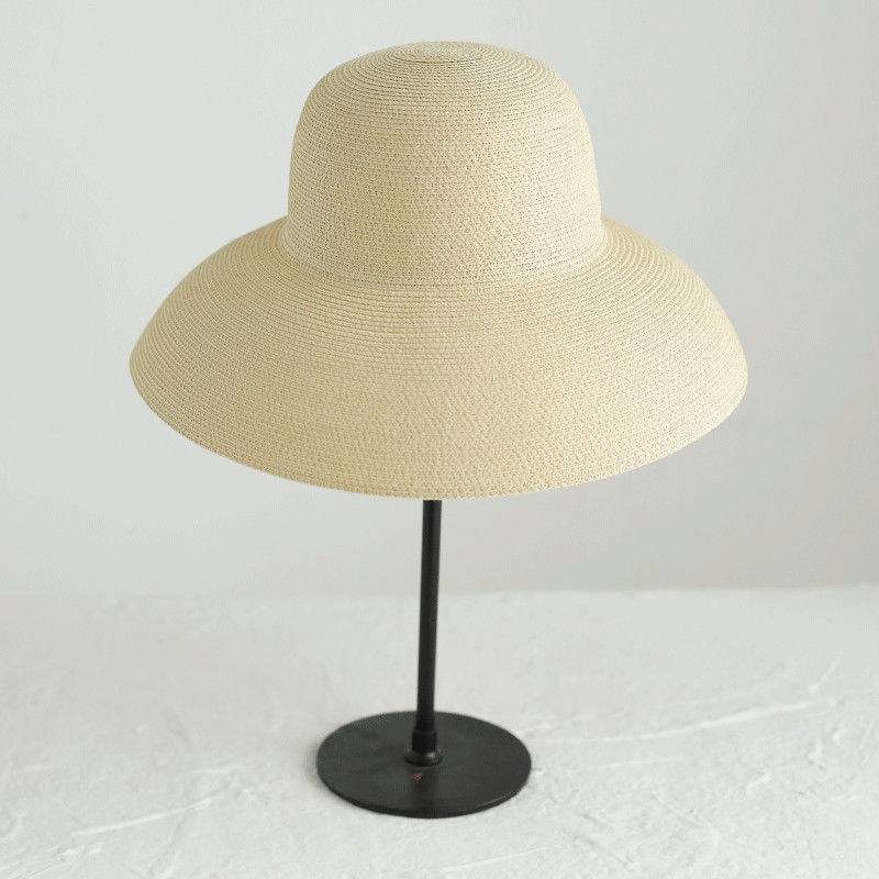 Chapeau bob beige en paille à large bord retombant posé sur un porte-chapeau noir avec un socle rond, le tout posé sur une table blanche, avec un mur blanc en fond