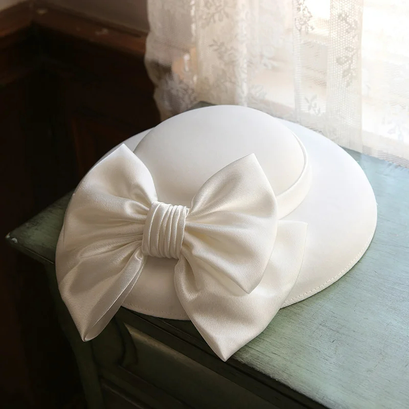 Chapeau mariage blanc style anglais avec un gros nœud, posé sur un meuble près d'une fenêtre.